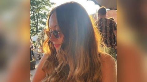 Stephanie Blundell, 41, poses wearing glasses while at an outdoor event. She has long brown hair and is smiling while looking to her right. 