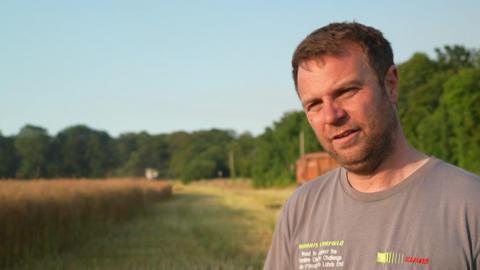 Farmer Olly Harrison with short brown hair and stubble wearing a beige t-shirt. He is standing in a field with trees behind him on a sunny day.
