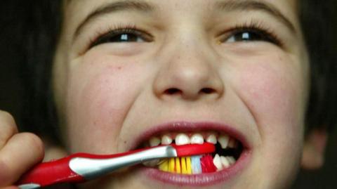 A close up of the face of a young child who is brushing their teeth with a red and white toothbrush which has red white and yellow bristles