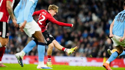 Exeter City player George Birch strikes a ball with his left foot during an FA Cup third round tie against Manchester City. He is wearing Exeter's home kit which is mostly red with black shorts and red socks. Birch has yellow boots on. Manchester City players are wearing sky blue shirts with white shorts and sky blue socks.