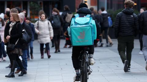 A Deliveroo rider, seen from behind, in a pedestrianised area. There are people walking either side of him. He is carrying a big Deliveroo branded backpack which is turquoise.