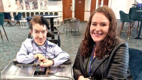 George Baker and Lauren Toovey smile at the camera. George is on the left in a powered wheelchair, sitting beside Lauren who is wearing a leather jacket in a spacious lounge area with chairs and tables in the background