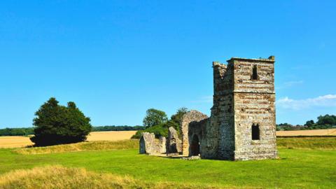 A derelict church sits in a mowed field with a tree to the left and blue skies above