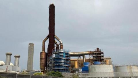 The Guernsey Electricity power plant is in the foreground, with a brown spiral shape and grey cylinders below it. A grey sky is in the background