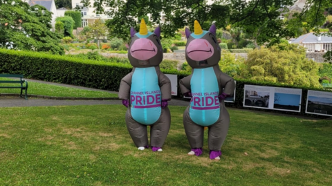 Two inflatable donkey unicorns, with a blue belly, grey skin, pink nose smiling and a yellow unicorn horn, on the stomach saying 'CHANNEL ISLANDS PRIDE' in a garden under a tree in the shade on a sunny day