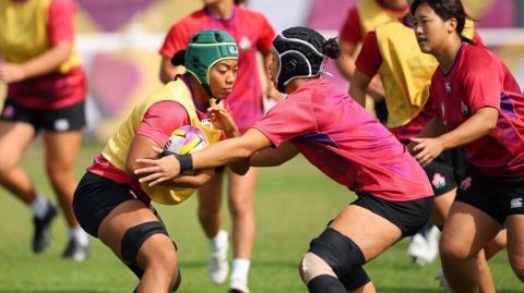 Women playing rugby, dressed in pink tops and black shorts. The player holding the ball is wearing a yellow bib and green helmet.  