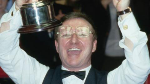 Dennis Taylor holding the trophy above his head in 1985. He is wearing glasses and a black bow tie and smiling, looking at the camera.