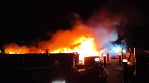 Fire in a marina at night.  Flames are visible surrounded by smoke.  A painted narrowboat name sign is visible to the right. A boat's chimney is visible to the left. 