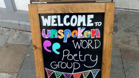 A blackboard with the words 'Welcome to Unspoken Word Poetry Group' written on it. The word unspoken has been written in colourful letters.