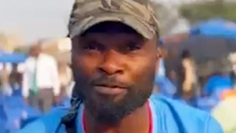 A DR Congo fan in a blue football shirt speaking to the camera in a crowd in Kinshasa