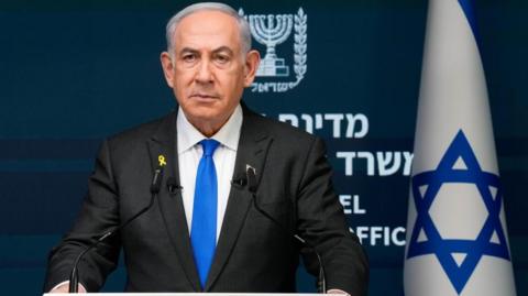 Israeli Prime Minister Benjamin Netanyahu speaking from a podium during a press conference. He is wearing a black suit and blue tie. Behind him is an Israeli flag. 