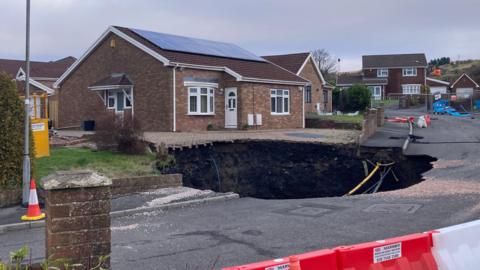 Large sinkhole in street on the drive of one house and into the road, with other homes nearby, barriers and cones are pictured in the street.