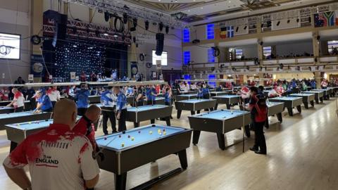 Pool players stand around dozens of pool tables in a large events space. The tables are covered in blue baize cloth and have red and yellow balls on them. The floor is of light polished wood. A stage area can be seen to the left. The flags of European nations hang from the rafters to the right.