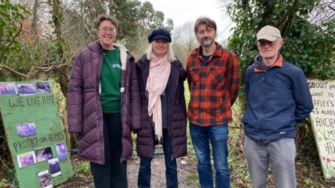Four people are standing, smiling at the camera. There are two women and two men. All are dressed for a walk in nature. There are two signs on either side of them and a field behind.