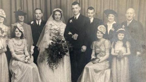 A black and white wedding photo. The bride is wearing a white dress and holding a large bouquet of flowers. She is linking arms with the groom who has a dark suit on. Three other men in suits are in the picture and there are several bridesmaids, including a girl with long dark hair. Another woman is wearing a jacket and dark hat. 