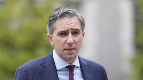 A man wears a navy suit and blue shirt with a maroon tie. He has short grey hair. Behind him, over his right shoulder is a out-of-focus green tree.  