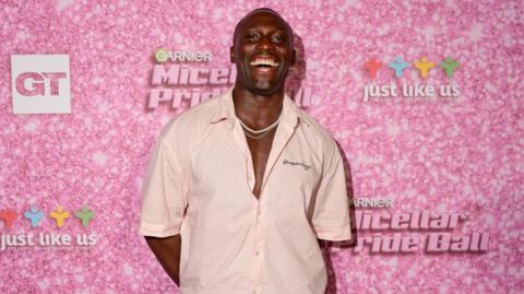 A man is pulling a big smile with his mouth open and is wearing a low-buttoned light pink shirt with some dainty white necklaces. Behind him is a pink wall which has the Garnier logo on it.