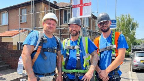 Three scaffolderts in harnesses stand in the sunshine outside a house which is covered in scaffolding. They are all wearing blue T-shirts. 