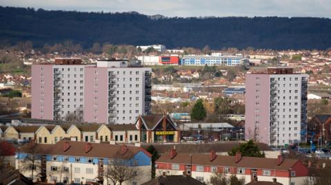 An aerial shot of Hartcliffe in south Bristol, with three of the area's distinctive pale-coloured tower blocks in the centre of the shot. A large Morrisons supermarket is also visible, and in the distance is a long ridgeline covered with trees