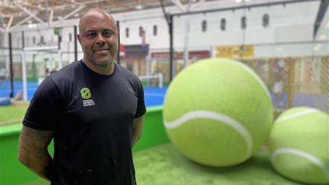 Image of Richard Belle who is stood in front of a padel court and two giant tennis balls