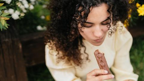 Stock photo of a close up shot of a person with black hair eating what looks like a chocolate covered protein bar outdoors