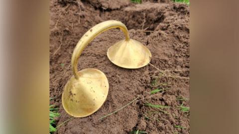 A curved gold dress fastener with conical ends is resting on a mound of dirt with tufts of grass popping through the mud.