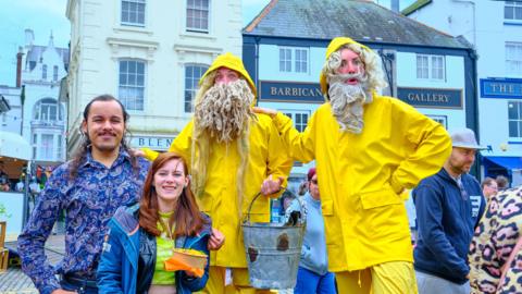 A photo which shows two people stood next to two other people wearing yellow fisherman outfits with fake white beards. One is holding a bucket.