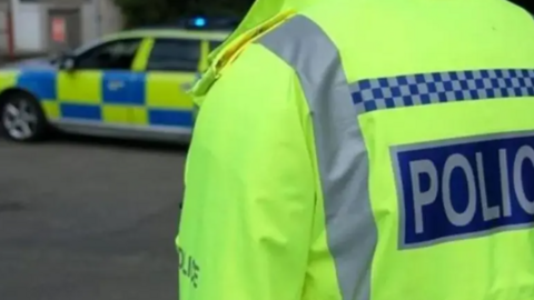 The back of a person wearing a high visibility police jacket. The words on the back of the jacket say 'POLICE' but the image is cropped before the end of the word. In the background, and slightly out of focus, is the side view of a police car.