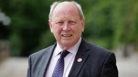 An older man looks into the camera smiling. he has grey hair at the side of his head and is bald on top. He is wearing a dark suit, white shirt and purple patterned tie. He is wearing a red badge that says 'no Irish sea border'.
