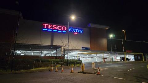 A Tesco Extra store with an illuminated sign and cones covering the entry road.