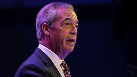 A side-view of Nigel Farage who is stood in front of a dark background. He is wearing a white shirt, dark suit and a tie.