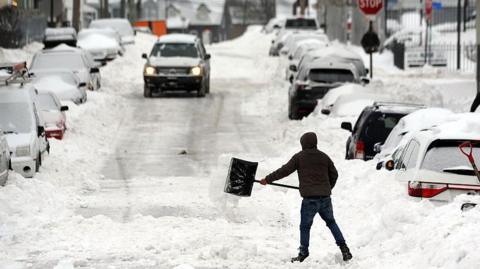 A man shovels out snow on a car lined road. Large amounts of snow surround the cars and a car is driving down the road.
