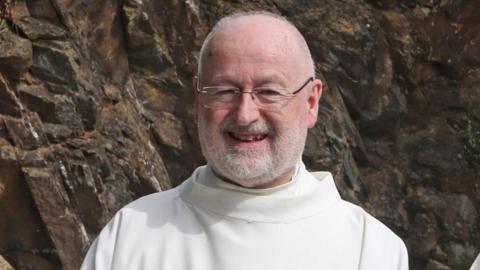 Fr John Murray. He is smiling at the camera. Fr Murray is bald and is wearing glasses. He has a white beard and is wearing white robes. Behind him is a rockface. 