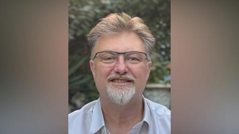 A man with glasses looking at the camera. He has light brown hair and a small beard which is grey. He is wearing a shirt and there is foliage behind him and possibly a large pot but it is a bit blurred.