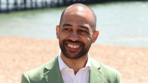 A bald man with a short beard smiling. He is wearing a light green suit and is standing on a beach with a pier behind him.