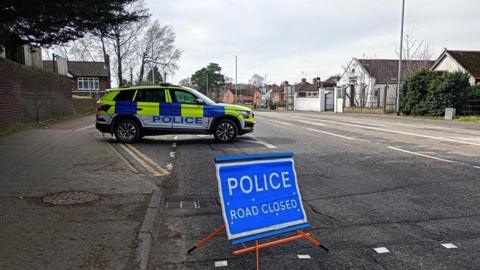 Scene in Dunmurry following a road traffic collision. There is a yellow, blue and white police vehicle to the left and a blue and white police road closed image