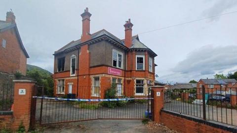 Gates are closed at the building where the fire broke out and police tape has been put up across the barrier. The two-storey building is made of brick and has chimneys. There is a shop sign on the front.