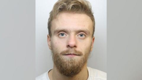 Mugshot of Ryan Scuffil, a 28-year-old man with pale skin, blue eyes, and blond hair. He has a blond moustache and short beard, and is wearing a white t-shirt.