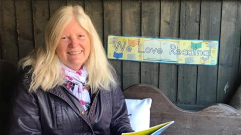 Paula Flood, wearing a brown leather jacket and a white and pink scarf, sits on a wooden bench. She is smiling at the camera. A sign behind her says "We love reading!"