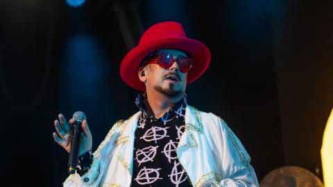 Boy George, wearing a red hat and grey shades, holds a microphone as he performs at the Henley Festival.