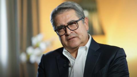 Close up of former Honduran President Juan Orlando Hernandez Alvarado in front of a yellow background. He is wearing a white shirt and black suit 