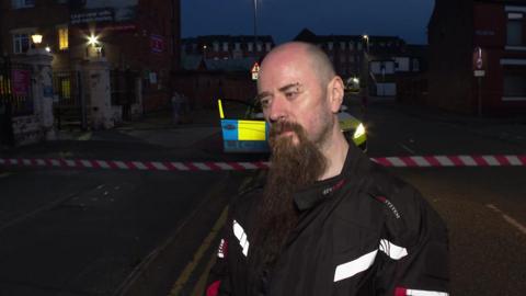 A bald man with a long beard and an eyebrow piercing, wearing a motorbike style jacket stands in a road that is closed off with tape. It is dark and a police car is behind him. 