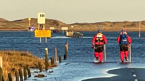 Two rescue officers in red dry suits wading into the sea. A partially submerged car can be seen in the distance as well as the white refuge hut.