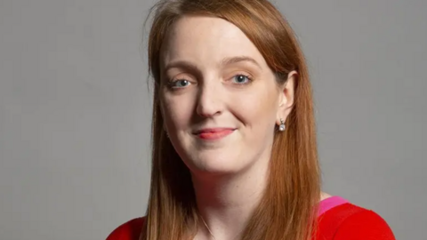 Portrait of Charlotte Nichols - Warrington North MP - with long red/light brown hair wearing a red and hot pink top. She is smiling.