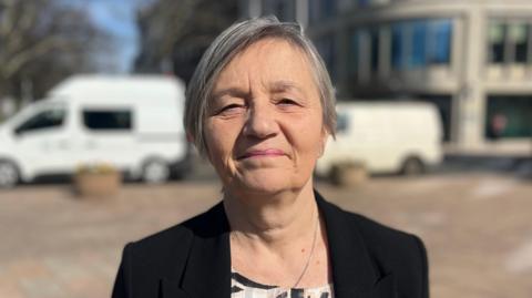 A head and shoulders picture of Deputy Mary Le Hegarat. She is wearing a black blazer and has short, grey hair. She is looking directly at the camera with a slight smile. The background is blurred but there is a building and two white vans in it.