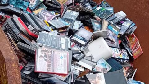 A photo of a skip filled with hundreds of DVDs. An electrical item is also in the skip.