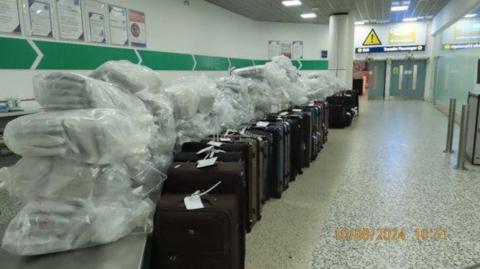 A long row of about 16 brown, blue and black suitcases are lined up with plastic bags dumped on the top of them containing white packets. The bags have been placed in an area of the airport with marble flooring and white walls with green arrows pointing towards a set of two large doors.