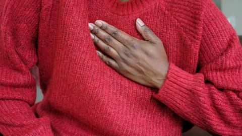 Black woman wearing a red knitted jumper placing a hand to her chest in pain.