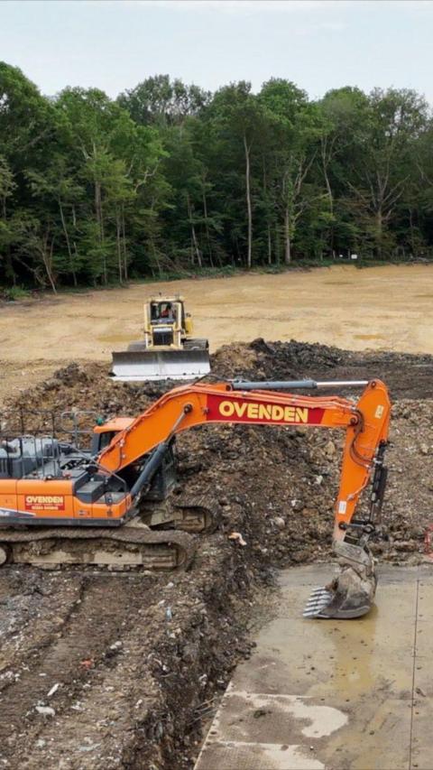 An orange digger digging up a clearing with a wood in the background