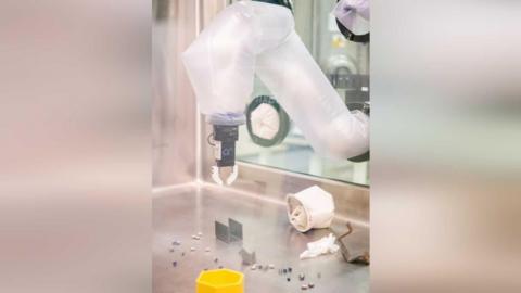 A robotic arm inside an active nuclear glovebox. The clear box has a a robotic arm with a flexible arm inside it. There are some metal objects inside the box, as well as a yellow hexagonal box.